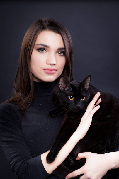 Portrait of a young brunette woman in black clothes on a black background with a black cat in her arms - Valokuva, kuva