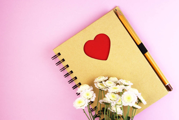 a brown notebook with flowers on a pink background. Notepad with a red heart and pencil on a pastel background. white camomile flowers at the bottom of the page - Photo, image