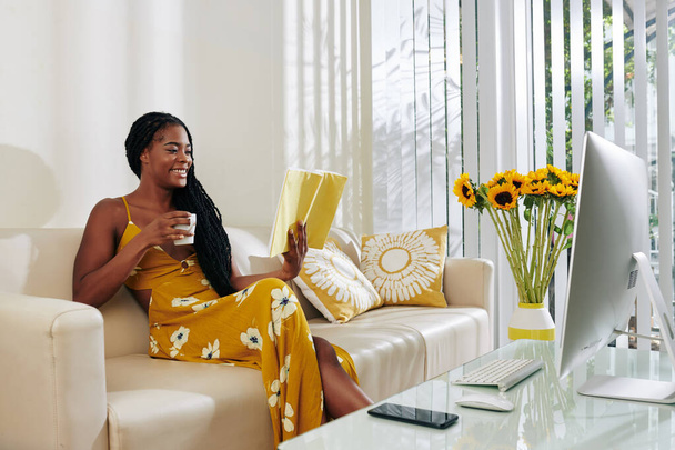 Όμορφη χαμογελαστή νεαρή γυναίκα πίνοντας φλιτζάνι νόστιμο καφέ και διαβάζοντας το βιβλίο όταν διαμένουν στο σπίτι το Σαββατοκύριακο - Φωτογραφία, εικόνα
