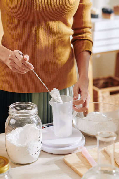 Обрезанное изображение женщины, помещающей щелочной порошок соды в пластиковую банку и измеряющей его в электронном масштабе при приготовлении мыльной смеси
 - Фото, изображение