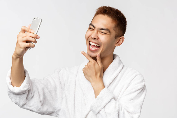 Koncepcja piękna, spa i technologii. Portret wesołego Azjaty w szlafroku spędzają covid19 kwarantanny na odległość społecznej w domu, robiąc selfie, patrząc uroczo na aparat komórkowy - Zdjęcie, obraz