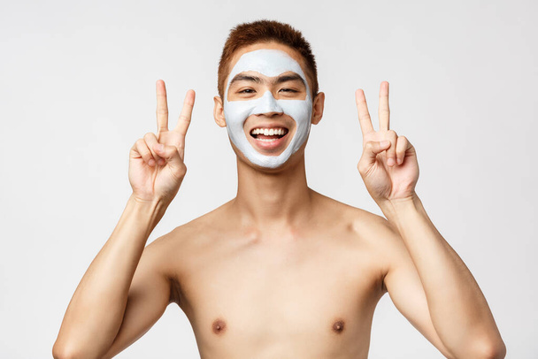 Krása, péče o pleť a lázeňský koncept. Veselý mladý Asiat s nahým trupem aplikované kožní rutinní kosmetiky, obličejová maska, ukázat mírové příznaky, relaxace doma, self-distancing, bílé pozadí - Fotografie, Obrázek