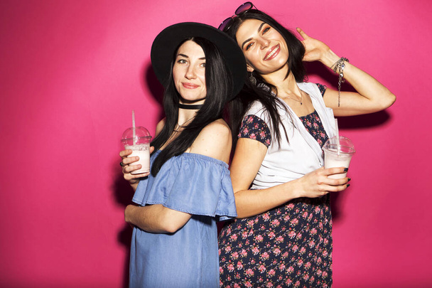 Deux brunes caucasiennes hipster femme en tenue décontractée élégante s'amuser à boire des milkshakes avec des pailles. Ils se tiennent sur un fond rose vif. Des émotions joyeuses et heureuses
 - Photo, image
