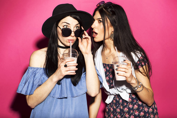 Twee Kaukasische brunette hipster vrouw in casual stijlvolle outfit met plezier drinken milkshakes met rietjes. Ze staan op een felroze achtergrond. Vrolijke, gelukkige emoties - Foto, afbeelding