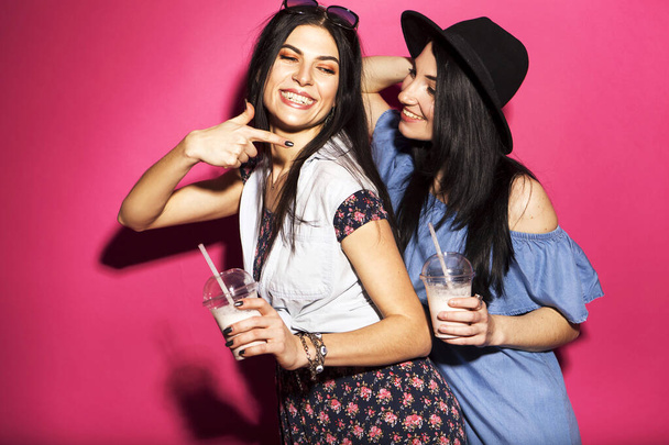Две кавказские брюнетки-хипстерши в повседневной стильной одежде, веселятся, выпивая молочные коктейли с соломинками. Они стоят на ярко-розовом фоне. Веселые, счастливые эмоции
 - Фото, изображение