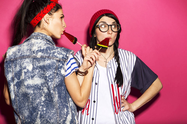 Két kaukázusi barna hipster nő alkalmi elegáns ruhában szórakozás nyalás görögdinnye nyalóka és játszik velük. Élénk rózsaszín háttéren állnak. Vidám, boldog érzelmek. - Fotó, kép