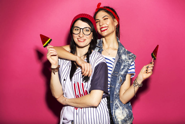 Dwie białe brunetka hipster kobieta w casual stylowy strój o zabawy lizanie lizaki arbuz lizaki i zabawy z nimi. Stoją na jasnoróżowym tle. Radosne, szczęśliwe emocje - Zdjęcie, obraz