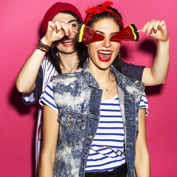 Twee Kaukasische brunette hipster vrouw in casual stijlvolle outfit met plezier likken watermeloen lolly 's en spelen met hen. Ze staan op een felroze achtergrond. Vrolijke, gelukkige emoties - Foto, afbeelding