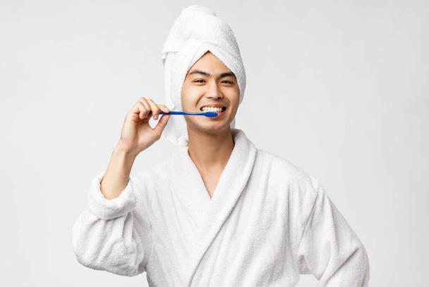 Концепция красоты, spa и отдыха. Веселый азиатский мужчина в халате и полотенце, чистит зубы зубной щеткой и улыбается, заботится о личной гигиене, белом фоне
 - Фото, изображение