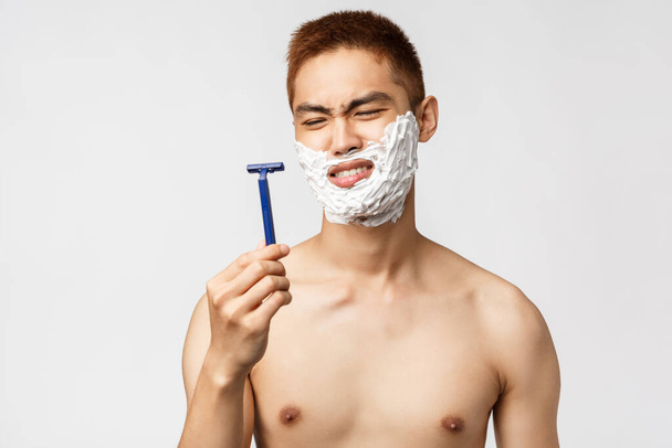 Beauty, people and hygiene concept. Portrait of disappointed, skeptical asian man grimacing, looking at razor displeased as it bad, shaving with old blades, apply facial cream to get rid of moustache - Zdjęcie, obraz