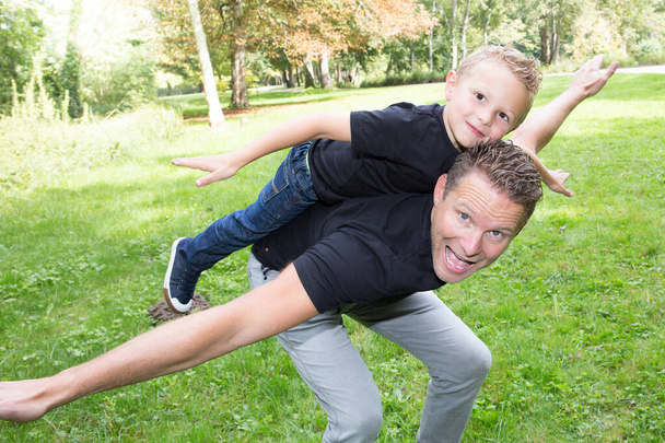 pohledný otec a roztomilý syn hrát zábava venku piggyback letadlo v zelené zahradě domů - Fotografie, Obrázek