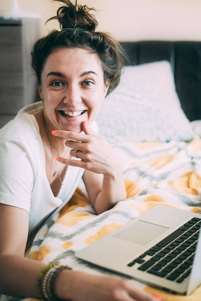 Ευτυχισμένη νεαρή γυναίκα κάθεται με ένα φορητό υπολογιστή στο κρεβάτι στο σπίτι, κοιτάζοντας την κάμερα και χαμογελώντας. Καλά νέα καραντίνας. Εύκολη μελέτη σε απευθείας σύνδεση. Επιτυχημένη δουλειά. Χαρούμενη επικοινωνία. Κλείσε. - Φωτογραφία, εικόνα
