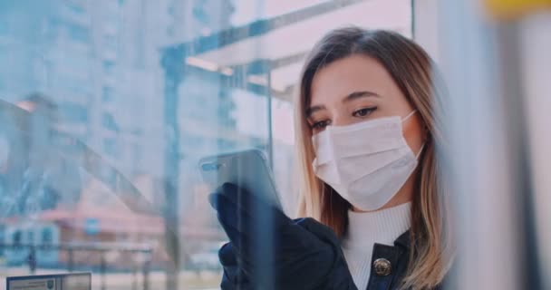 Close up portrait of young smart female volunteer in a medical mask is texting on the phone at bus stop. Quarantine COVID-19 in Europe. Coronavirus transmission in public transport. - Πλάνα, βίντεο
