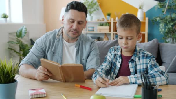 Caring dad reading book to kid while child drawing pictures at desk in apartment - Metraje, vídeo