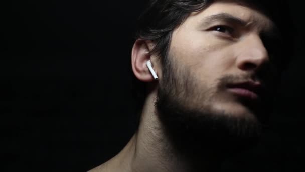 Dunkles Nahaufnahme-Porträt eines jungen Mannes mit drahtlosen Kopfhörern auf schwarzem Hintergrund. - Filmmaterial, Video