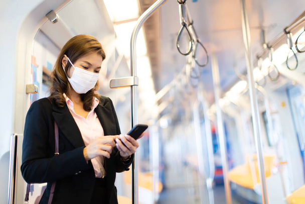Bueatiful adult Asian businesswoman passenger commuter wearing medical hygiene protect mask. Standing and using cell phone inside empty subway or sky train car. Idea for abandoned city from COVID-19. - Foto, Imagem
