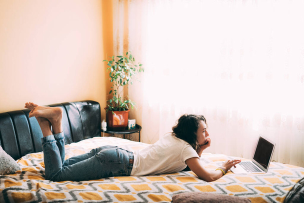 A young girl with curly hair in a white T-shirt and jeans lies on the bed and looks at the laptop. Full size side view. Sunny room. Stay home concept. Work and study online. Copyspace. Soft focus - Photo, Image