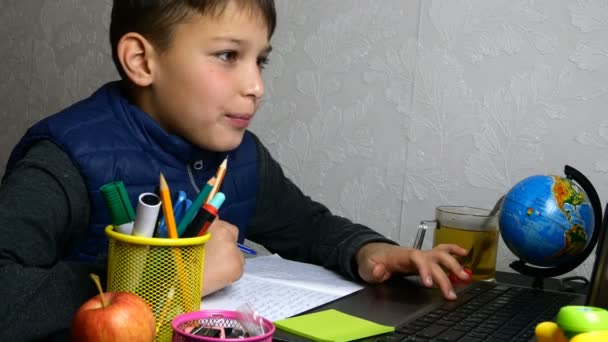 Διδασκαλία εξ αποστάσεως, ηλεκτρονική μάθηση. Schoolboy παίρνει online εκπαίδευση και κάνει σχολική εργασία κατά τη διάρκεια της καραντίνας με σημειωματάριο. Καθισμένος στην επιφάνεια εργασίας με σχολικά είδη. Κοντινό πορτραίτο. - Πλάνα, βίντεο