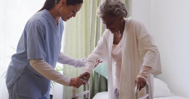 A Caucasian nurse wearing blue scrubs, helping a senior African American woman walking with a walking frame, in a retirement home, during coronavirus covid19 pandemic. - Кадры, видео