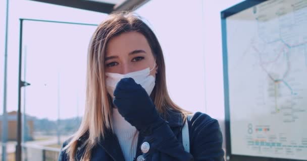 portrait of young cute girl wearing in medical mask staying and waiting for bus at a public transport stop and looking to camera. Concept health and safety, coronavirus quarantine, virus protection - Кадри, відео