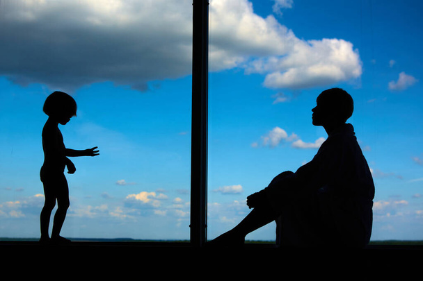 Anya sziluettjei 3 éves lányával egy nagy ablak előtt, kék égbolttal és felhőkkel. Maradj otthon a koronavírus-világjárvány karantén alatt tartására. Önizolálás - Fotó, kép