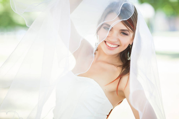 Close up of attractive young woman in elegant veil looking at camera and smiling. Charming newlywed in white dress rejoicing and demonstrating charming smile. Concept of bride beauty and happiness. - Fotó, kép
