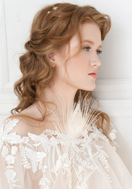 Beautiful natural redhead girl bride, with nude makeup, wearing a white dress, stands at the white wall in light interior. Close up portrait - 写真・画像