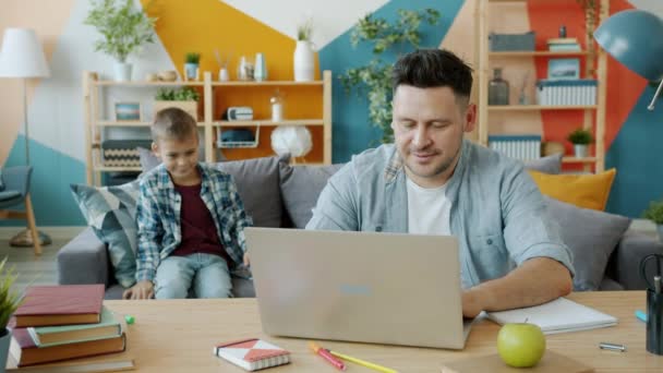 Dad using computer sitting at desk while son approaching him talking at home - Filmmaterial, Video