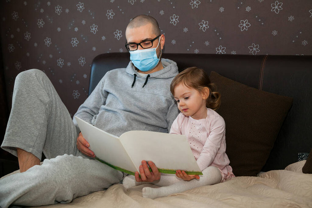 Father in protective mask spending time together with his daughter during quarantine time because of coronavirus outbreak. They are reading the book. - Foto, afbeelding