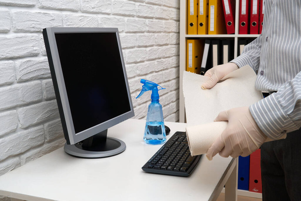 Konzept der Reinigung oder Desinfektion des Büros - ein Geschäftsmann reinigt den Arbeitsplatz, Computer, Schreibtisch, verwendet eine Spritzpistole und Papierservietten. Reinigung von Oberflächen von Mikroben, Viren und Schmutz. - Foto, Bild