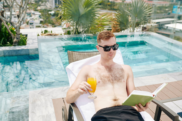 Σοβαρός νεαρός άνδρας με γυαλιά ηλίου πίνει αναζωογονητικό χυμό πορτοκαλιού και διαβάζει ενδιαφέρον βιβλίο όταν ξεκουράζεται δίπλα στην πισίνα - Φωτογραφία, εικόνα