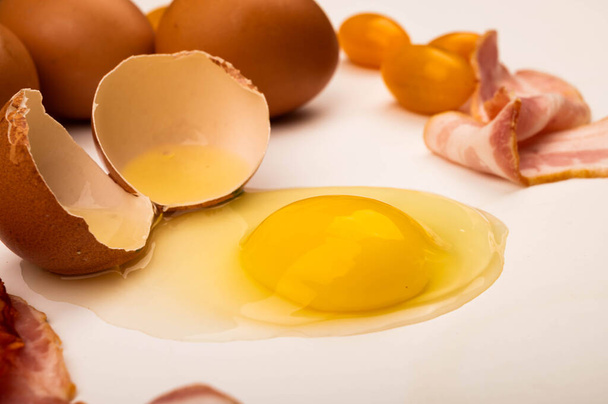 Ovo de galinha quebrado, ovos de galinha, fatias de bacon e tomates em um fundo branco. Fechar
 - Foto, Imagem