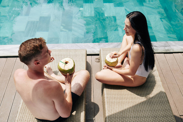 Joven pareja sonriente sentada en chaise-lounges junto a la piscina en un día soleado, bebiendo cócteles de coco y hablando
 - Foto, Imagen