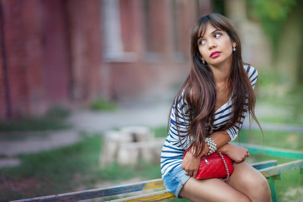 Front view of beautiful lady with long hair holding elegant red purse and looking aside while sitting on bench outdoors. Charming girl in stylish striped shirt with blurred background and copy space. - Foto, immagini