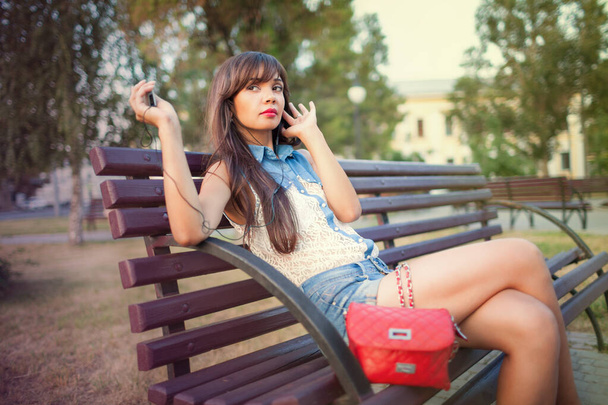 Side view of beautiful girl with long hair listening to music on mp3 player and singing. Charming lady sitting on bench and enjoying favorite songs. Concept of love for music. - Foto, immagini