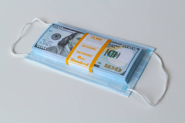 orvosi maszk és egy halom 100 dolláros bankjegy a magasabb árak szimbólumaként és a légutak vírusokkal szembeni védelmének hiányaként. A védőmaszkok árának emelkedése. - Fotó, kép