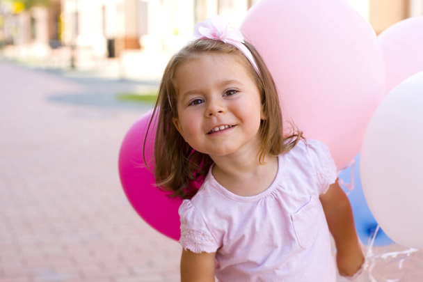 όμορφο μελαχρινό μωρό με ροζ φόρεμα και με ένα ροζ λουλούδι στο κεφάλι της περπατάει στο δρόμο με μπαλόνια στο χέρι και χαμογελάει χαριτωμένα στην κάμερα - Φωτογραφία, εικόνα