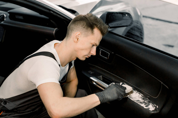 Pohledný mladý muž, pracovník profesionální autodílny, mytí interiéru dveří auta měkkým štětcem s pěnou. Úklid a detailní koncepce - Fotografie, Obrázek