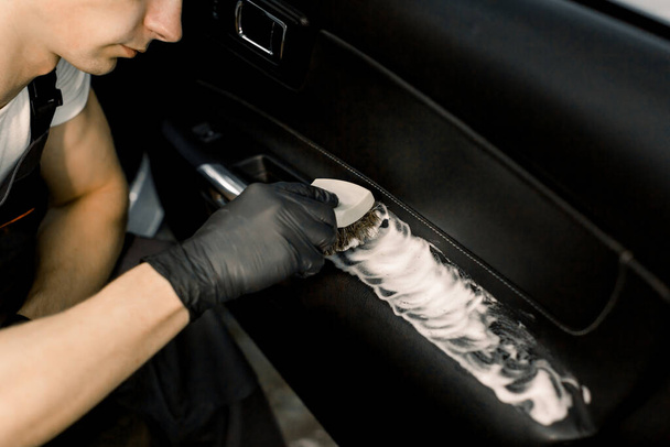 Cropped image of washing car interior by a soft brush with foam. Young male worker in black protective gloves cleaning car door inside with foam and brush - Фото, изображение