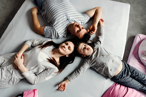 Top view of a cute little girl and her beautiful young parents looking at the camera and smiling, lying on the floor at home,Vvew from above of resting family lying on the floor and looking at camera - Fotografie, Obrázek