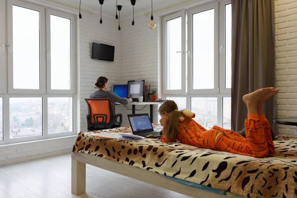 Семья дома, ребенок лежит на кровати и учится в ноутбуке, мать на заднем плане работает дистанционно в компьютере
 - Фото, изображение