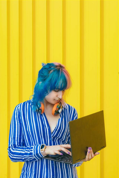 A young female with pink and blue hair wearing a blue striped shirt holding a laptop against a yellow background - Φωτογραφία, εικόνα