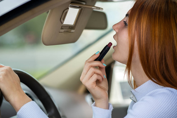 Detailní záběr mladé zrzky žena řidič opravuje její make-up s tmavě červenou rtěnkou při pohledu do auta zpětné zrcátko za volantem vozidla. - Fotografie, Obrázek