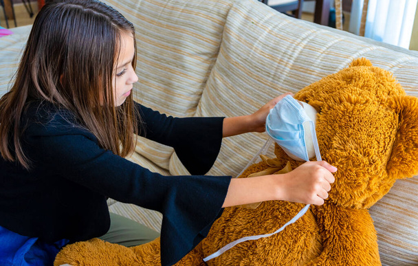 Hermosa niña en una chaqueta negra jugando con su oso de peluche marrón que ha puesto una máscara para protegerlo del coronavirus en el sofá blanco en casa. concepto de inocencia
 - Foto, imagen