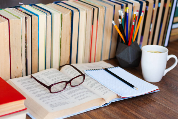 Open textbook, notebook, glasses, pencils in holder, cup of tea and stack of old book on wooden table, education concept background, many books piles with copy space for text - Foto, Bild