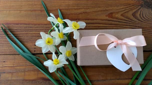 白い水仙と茶色いギフトボックス、木製の背景4Kビデオでピンクリボン付きグリーティングカード。女性の手は休日現在のパッケージの周りに花を置きます。3月8日ハッピーレディースマザーズバレンタインデー - 映像、動画