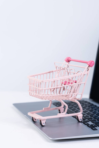 Online shopping. Mini empty pink shop cart trolley over a laptop computer on white table background, buying at home concept, close up - Photo, Image