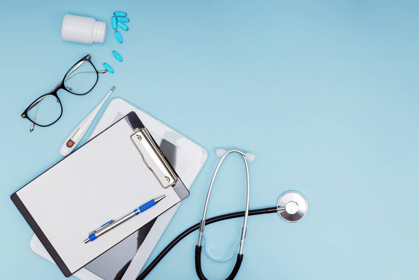 Workplace of modern doctor concept. Tablet, glasses, pills, stethoscope and other objects on blue background - Photo, image