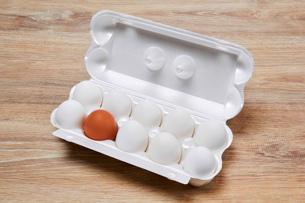 nove ovos brancos e um marrom são embalados juntos em uma caixa de ovo em uma superfície de mesa de madeira
 - Foto, Imagem
