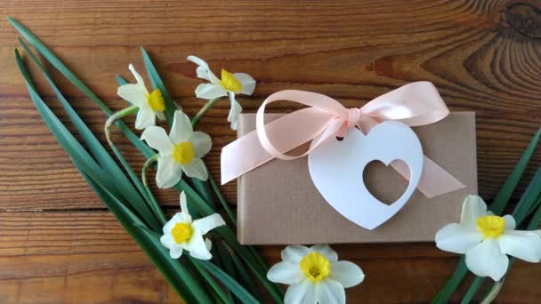 Białe żonkile i brązowe pudełko z prezentami, kartka z różową wstążką na drewnianym tle wideo 4K.Żeńska ręka kładzie kwiaty wokół obecnych na wakacjach opakowań. 8 marca Szczęśliwych Kobiet Matki Walentynki - Materiał filmowy, wideo